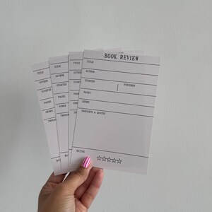 May include: A stack of white rectangular book review cards featuring black printed text. Each card includes fields for Title, Author, Started, Finished, Pages, Genre, Thoughts & Quotes, and a Rating section with five empty star outlines at the bottom.