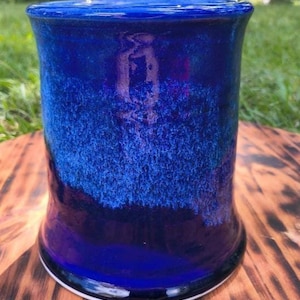 May include: A handmade ceramic mug with a deep blue glaze. The mug has a cylindrical shape with a slightly flared base and a smooth, glossy finish. The color transitions from a lighter blue at the top to a darker shade at the bottom. The mug is approximately 5 inches tall.