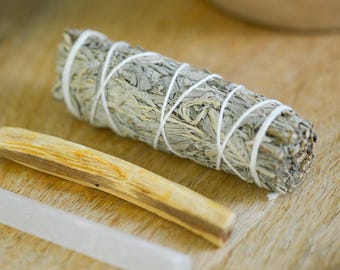 May include: A bundle of dried sage wrapped with white string, a piece of palo santo wood, and a clear selenite wand. The items are arranged on a light brown wooden tray, suggesting a focus on natural wellness or spiritual practices.