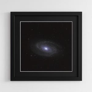 May include: A framed print featuring a spiral galaxy with a bright, blue-white core and swirling arms against a dark, star-filled background. The print is set within a black frame with a white mat.