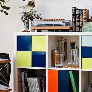May include: A white cube shelf unit with colorful accents. The unit features blue and yellow squares, books, a record player, and decorative items. A framed artwork hangs above the shelf, adding to the room's decor.