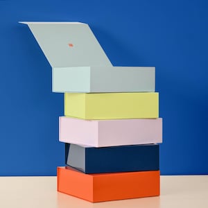 May include: A stack of six colorful rectangular boxes against a blue background. The boxes are in shades of orange, navy, pink, yellow, and light blue. One box is open, revealing a small red square inside.
