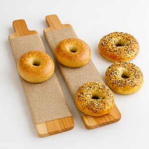 May include: Wooden serving boards with a natural burlap wrap, each holding a bagel. Two plain bagels and three everything bagels are also displayed. The bagels are golden brown with a variety of toppings.