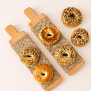 May include: Overhead shot of two wooden serving boards with burlap wraps, each holding a bagel. Four additional bagels are scattered around. The bagels have various toppings, including sesame seeds and everything seasoning.