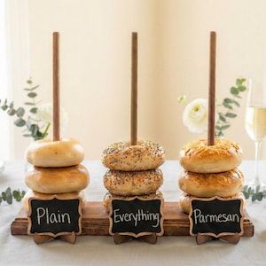 May include: Three wooden bagel stands with bagels labeled "Plain", "Everything", and "Parmesan". Each stand has a wooden dowel through the center of the bagels. The stands are on a wooden base with small chalkboard signs.
