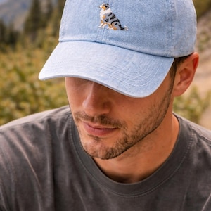 May include: Light blue baseball cap with a curved brim. The cap features an embroidered image of a dog in shades of black, white, and brown. The cap is made of a soft, textured fabric.