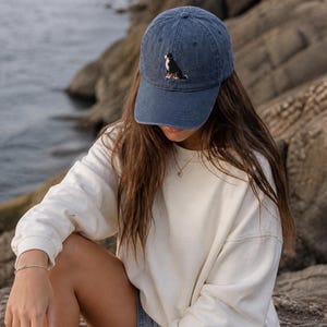 May include: A navy blue baseball cap with an embroidered Bernese Mountain Dog design. The cap has a curved brim and a classic six-panel construction. The dog is depicted in black, white, and brown. The cap is made of a soft, washed cotton material.