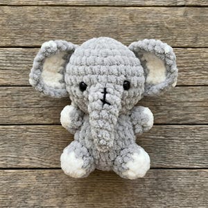 Puede incluir: Un peluche de elefante gris hecho a mano con ganchillo, con detalles blancos en las orejas y los pies. El elefante tiene ojos de seguridad negros y una trompa cosida. El juguete suave está diseñado para niños.