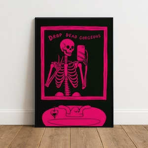Könnte beinhalten: Ein gerahmter Kunstdruck mit einem rosa Skelett, das ein Selfie macht. Das Skelett befindet sich in einem rosa Rahmen mit den Worten "DROP DEAD GORGEOUS" oben. Der Hintergrund ist schwarz, und der Druck wird an einer Wand angezeigt.