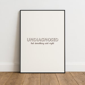 Może przedstawiać: Oprawiony wydruk artystyczny z napisem "UNDIAGNOSED but something ain't right" w kroju pisma szeryfowego. Wydruk ma kremowe tło i czarną ramę. Grafika jest eksponowana na drewnianej podłodze na tle białej ściany.