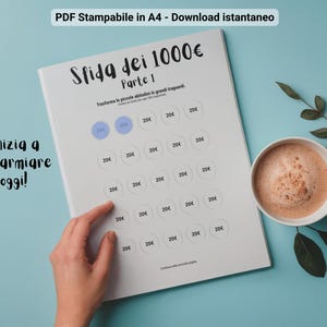 Op de afbeelding: Een witte, afdrukbare spaaruitdagingsvel met de tekst "Sfida dei 1000€ Parte 1" en "Inizia a risparmiare oggi!" wordt weergegeven op een lichtblauwe ondergrond. Een kopje koffie staat naast het vel.