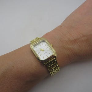 Peut inclure: Une montre à quartz dorée avec un cadran rectangulaire et un bracelet à maillons dorés. Le cadran de la montre est blanc avec des chiffres romains noirs et des aiguilles dorées. Le mot "Quartz" est imprimé sur le cadran.