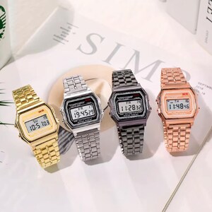 Puede incluir: Cuatro relojes de pulsera digitales con esferas rectangulares y correas de eslabones metálicos. Los relojes tienen acabados en oro, plata, gris oscuro y oro rosa. Cada reloj muestra la hora, el día y la fecha, con el texto WR en la esfera.