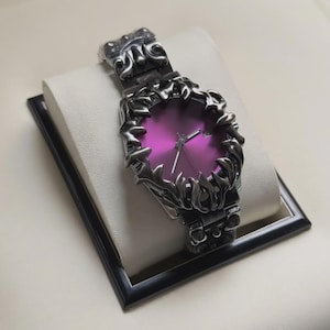 Puede incluir: Reloj de pulsera plateado con esfera morada vibrante. El reloj presenta un bisel y una correa ornamentados y estilizados. El reloj se exhibe sobre un cojín blanco dentro de un marco negro.