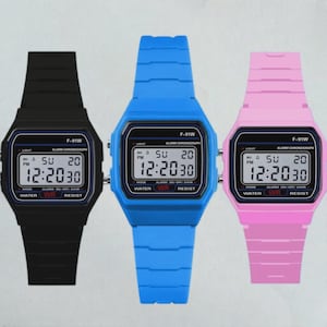Peut inclure: Trois montres numériques en noir, bleu vif et rose clair. Chaque montre présente un cadran rectangulaire avec un affichage numérique noir indiquant l'heure 12:20, la date, et le texte F-91W, ALARM CHRONOGRAPH, et WATER WR RESIST.