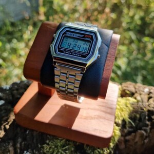 May include: A silver digital watch with a black face and a stainless steel band is displayed on a wooden stand. The watch shows the time as 9:29:88. The stand is made of wood and has a black leather cushion.