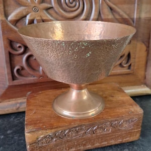 May include: A hammered copper bowl with a wide, flared rim and a pedestal base. The exterior surface features a textured, dimpled finish with visible green oxidation spots scattered across the metallic body, showing a warm, aged copper tone.