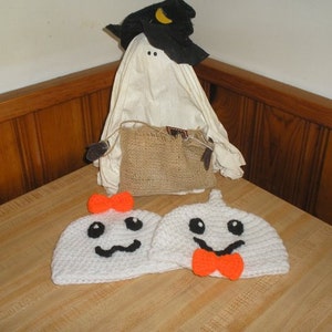 May include: Two white crocheted ghost hats with black eyes and mouths, one with an orange bow and the other with a red bow. The hats are sitting on a wooden surface.