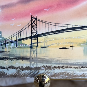 May include: A watercolor painting depicting a suspension bridge over a body of water at sunset. The sky features shades of pink and orange, while the water reflects the bridge and distant city silhouettes. Small boats are visible on the water.