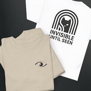 May include: Two t-shirts, one white and one beige. The white shirt features a black cat graphic inside an arch with the text "INVISIBLE UNTIL SEEN." The beige shirt has a small eye graphic. Both are folded.