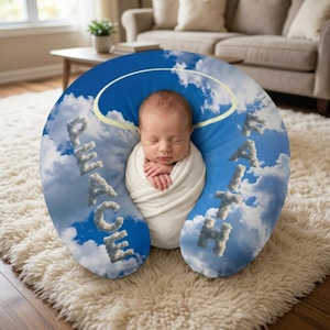 Puede incluir: Una almohada de lactancia en forma de U con un estampado de cielo azul y nubes blancas. La palabra PEACE está escrita en el lado izquierdo y FAITH en el derecho, ambas formadas por nubes. Un gráfico de halo amarillo se sitúa arriba.