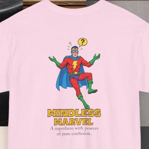Könnte beinhalten: Hellrosa T-Shirt mit einem Comic-Superhelden-Design. Der Superheld in einem rot-blauen Kostüm hat ein Fragezeichen über dem Kopf. Der Text "MINDLESS MARVEL" und ein Slogan sind unter dem Bild des Superhelden aufgedruckt.