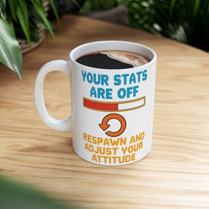 May include: White ceramic coffee mug with black coffee. The mug features the text "YOUR STATS ARE OFF" in blue, a red and orange loading bar, and the text "RESPAWN AND ADJUST YOUR ATTITUDE" in orange.