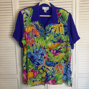 Puede incluir: Una camisa de manga corta con botones y un vibrante estampado floral. La camisa presenta un cuello y mangas azules, con un diseño colorido de hojas y flores en tonos verdes, naranjas, rosas y blancos. La camisa está colgada en una percha blanca.