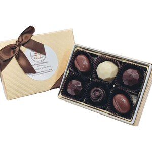 Op de afbeelding: Een gouden doos met een bruin lint en een label met de tekst "Artisan Bonbons 6 Piece Antique Collection" bevat zes verschillende chocoladebonbons. De bonbons zijn gerangschikt in een witte en gouden doos met een zwarte voering.