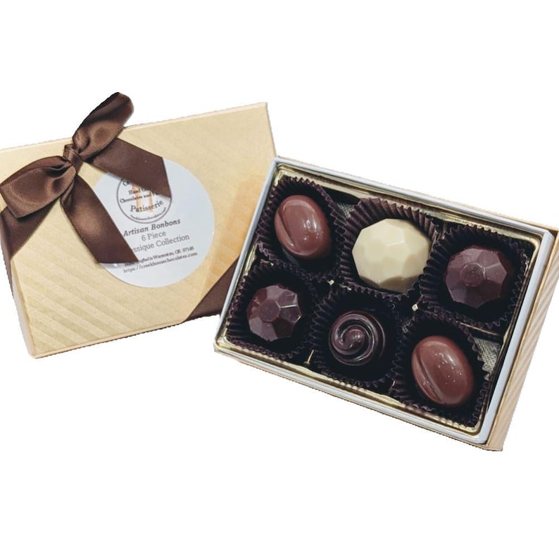 Op de afbeelding: Een gouden doos met een bruin lint en een label met de tekst "Artisan Bonbons 6 Piece Antique Collection" bevat zes verschillende chocoladebonbons. De bonbons zijn gerangschikt in een witte en gouden doos met een zwarte voering.