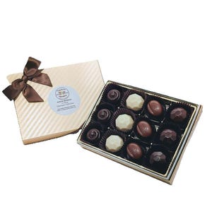 Op de afbeelding: Een doos met verschillende chocolaatjes in een gouden en witte gestreepte doos. De doos is open en de chocolaatjes zijn in een rasterpatroon gerangschikt. De chocolaatjes zijn donker, melk en wit.