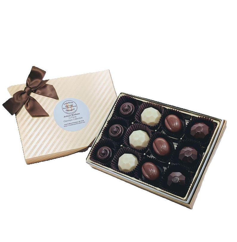 Op de afbeelding: Een doos met verschillende chocolaatjes in een gouden en witte gestreepte doos. De doos is open en de chocolaatjes zijn in een rasterpatroon gerangschikt. De chocolaatjes zijn donker, melk en wit.