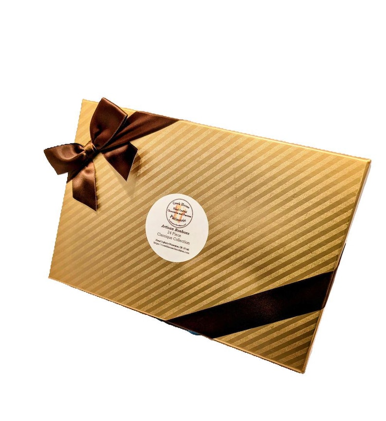 Op de afbeelding: Een gouden doos met een bruin lint en een wit rond label met de tekst "Artisan Bonbons 24 Stuks Classic Collection".