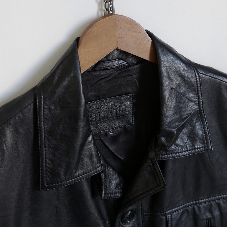 May include: Black leather jacket with a collared neckline and button closure. The jacket features a label that reads "SHATIS" and the number "52". The jacket is hanging on a wooden hanger.