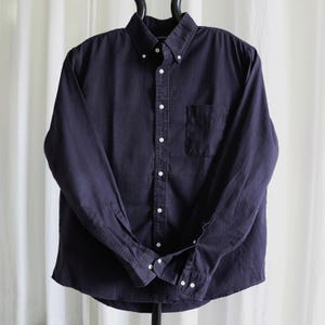May include: A dark navy blue button-down shirt with a button-down collar and a single chest pocket. The long-sleeved shirt has white buttons and is hanging on a black hanger. The shirt is made of a soft, durable fabric.