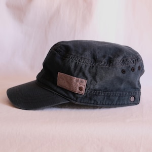 Puede incluir: Una gorra de estilo militar gris oscuro con una textura de tela de patrón de rejilla. El lateral de la gorra muestra un parche rectangular color canela con el texto 301, dos ojales de metal oscuro y un pequeño botón metálico en el borde.