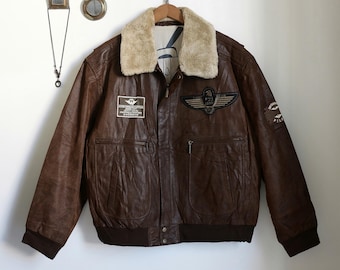 May include: A brown leather bomber jacket with a faux fur collar. The jacket features multiple patches and pockets, with a ribbed hem and cuffs. The jacket is hanging on a wooden hanger against a white wall.