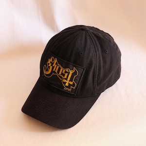Könnte beinhalten: Eine schwarze Baseballkappe mit einem rechteckigen Aufnäher auf der Vorderseite. Der Aufnäher zeigt das Wort Ghost in einer stilisierten goldenen Schrift mit einem in den Schriftzug integrierten Kreuz. Der Text 2015 GHOST steht in kleinen Buchstaben.
