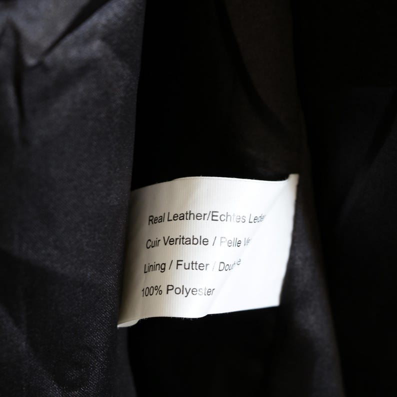 May include: Close-up of a black garment's interior, revealing a white label. The label displays text indicating "Real Leather," "Cuir Veritable," and "100% Polyester." The garment's lining is visible.