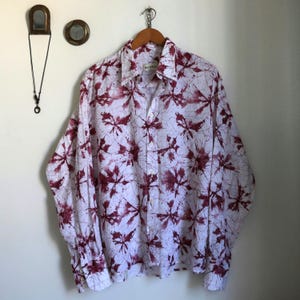 Peut inclure: Chemise blanche à manches longues avec un motif de feuilles marron. La chemise est suspendue à un cintre en bois. La chemise a un col et des boutons. La chemise est faite d'un tissu léger.