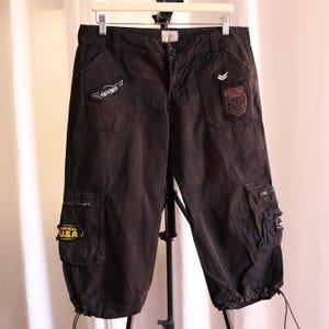 May include: Black cargo capri pants featuring multiple pockets, a button and zipper fly, and belt loops. The pants are decorated with various embroidered patches, including one that reads AIR FORCE, one that says MILITARY, and one stating MICHIGAN U.S.A.