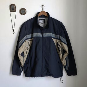 May include: Navy blue zip-up jacket with beige and gray accents. The jacket features a high collar, a logo on the chest, and the word "Quiksilver" in a stylized font on the sleeve. The jacket has a drawstring at the bottom.