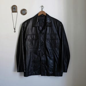 May include: A black leather jacket with a collared neckline and button-up front. The jacket has two chest pockets with buttoned flaps and is hanging on a wooden hanger. The jacket is a classic style.