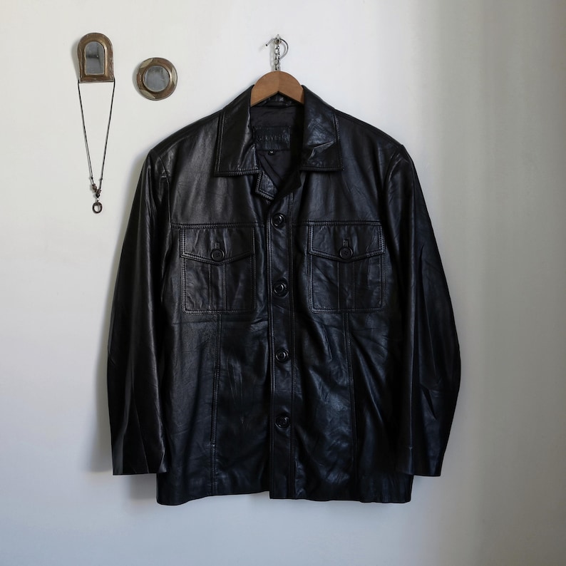 May include: A black leather jacket with a collared neckline and button-up front. The jacket has two chest pockets with buttoned flaps and is hanging on a wooden hanger. The jacket is a classic style.