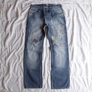 Puede incluir: Vaqueros de mezclilla azul claro con parches decorativos y detalles bordados. Los vaqueros tienen bragueta con botones y diseño clásico de cinco bolsillos. Los parches incluyen formas bordadas y pequeñas piedras decorativas.