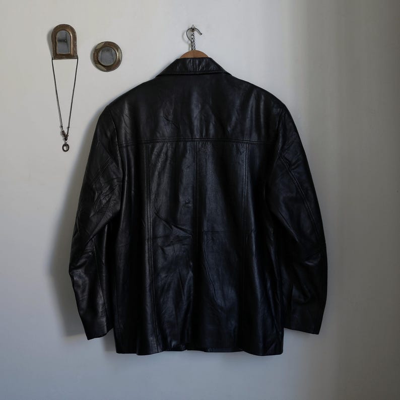 May include: A black leather jacket hangs on a wooden hanger against a white wall. The jacket has a classic design with a collar and button closure. Decorative items, including a small mirror and a necklace, are mounted on the wall to the left of the jacket.