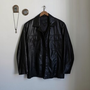 May include: A black leather jacket with a button-up front and two chest pockets. The jacket is hanging on a wooden hanger against a white wall. The jacket has a classic, vintage style.