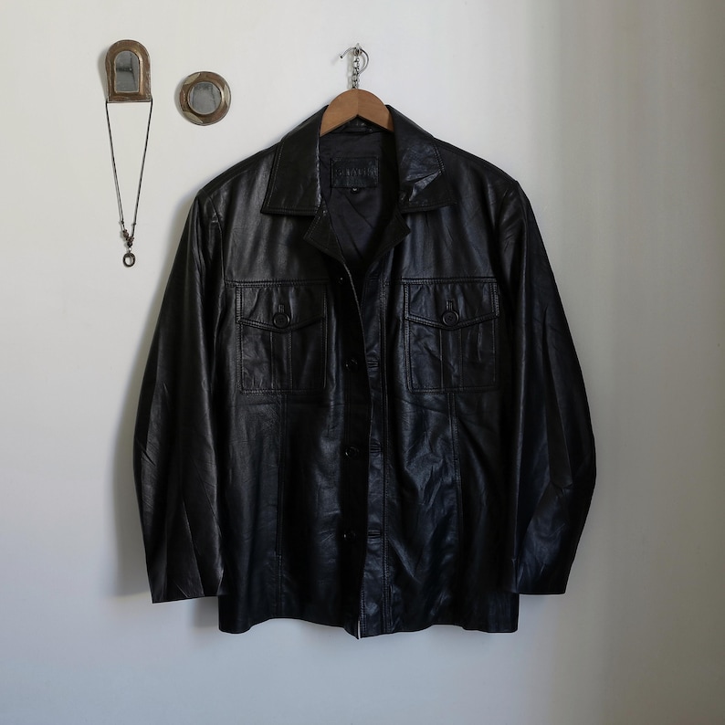 May include: A black leather jacket with a button-up front and two chest pockets. The jacket is hanging on a wooden hanger against a white wall. The jacket has a classic, vintage style.