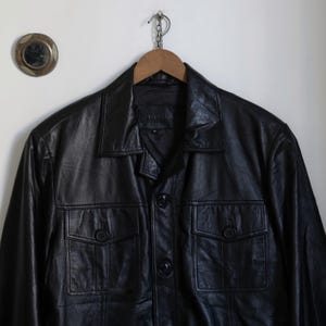 May include: A black leather jacket with a collared neckline and two front pockets. The jacket is buttoned up and hanging on a wooden hanger. The jacket has a classic, timeless design.