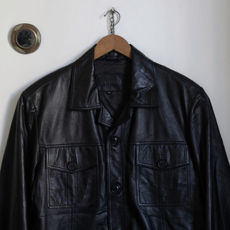 May include: A black leather jacket with a collared neckline and two front pockets. The jacket is buttoned up and hanging on a wooden hanger. The jacket has a classic, timeless design.